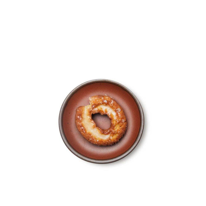 A donut on a dessert sized ceramic plate in a cool burnt terracotta color featuring iron speckles and an unglazed rim.