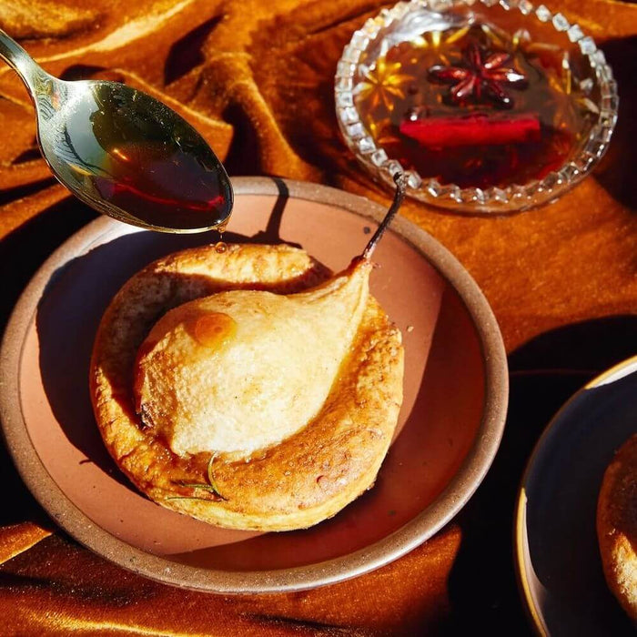 Spoon drizzling syrup over a baked pear dessert on a cool terracotta ceramic dessert plate with spiced syrup in the background.