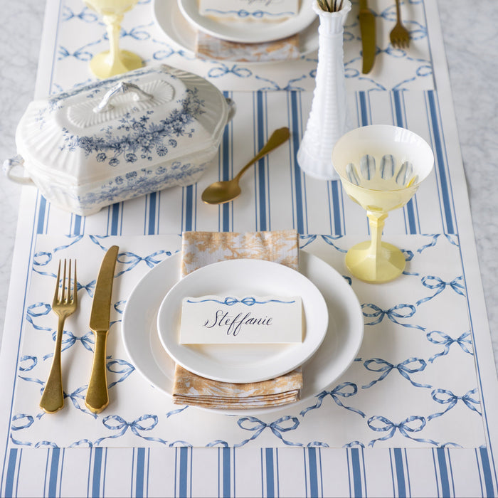 Elegant table setting with Heirloom Blue Bow Lattice Placemat, Heirloom Blue Bow Place Card, Cornflower Blue Linen Stripe Runner, white plates, gold cutlery, and decorative elements.