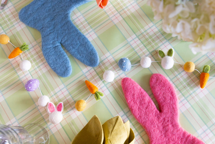 A close-up of an Easter themed table, featuring pink and blue felt bunny placemats, a felt garland, and a green plaid backdrop.