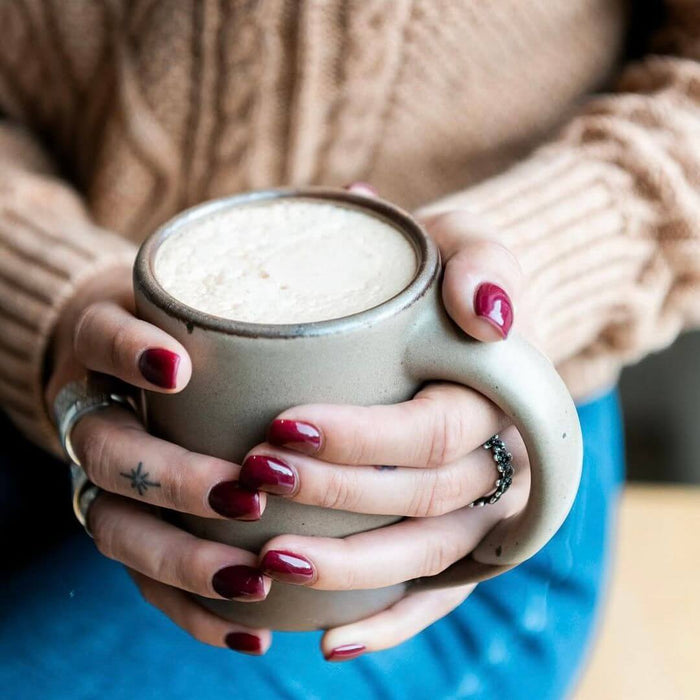Photo by @farmettemichigan. Hands wrapping around a medium sized ceramic mug with handle in a warm pale brown color featuring iron speckles.
